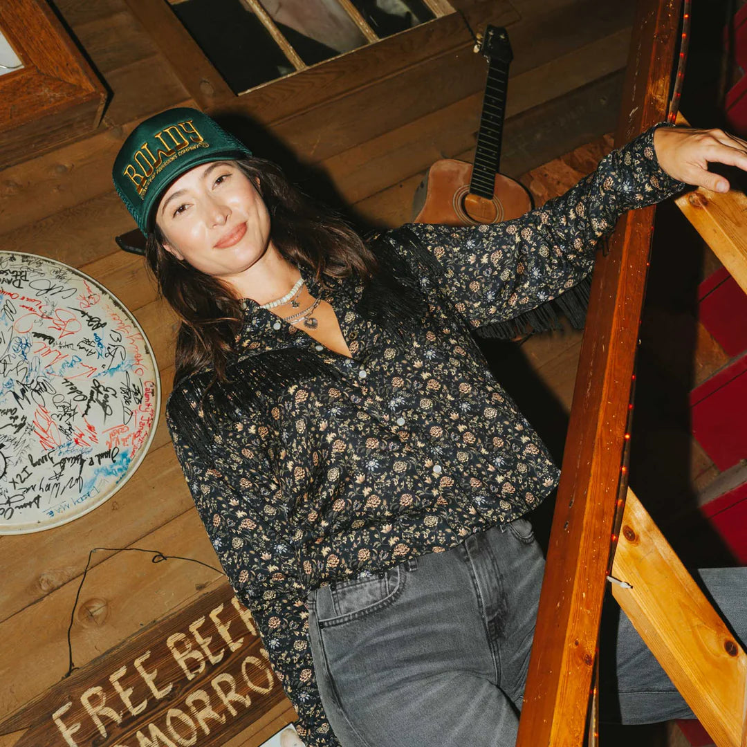 Woman wearing a floral shirt and green cap in a rustic setting with a guitar and wooden decor.