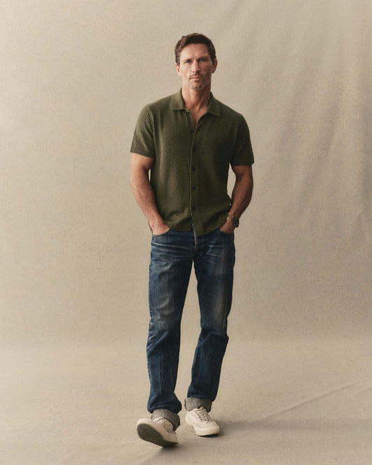 Man wearing a green shirt and blue jeans standing against a plain background
