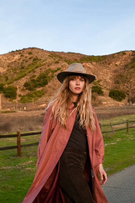 Woman wearing a long coat and hat in a scenic outdoor setting with mountains and a fence.