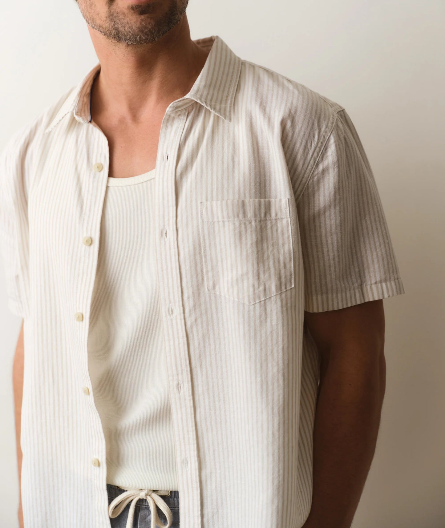 Man wearing a beige striped shirt over a white tank top against a plain background