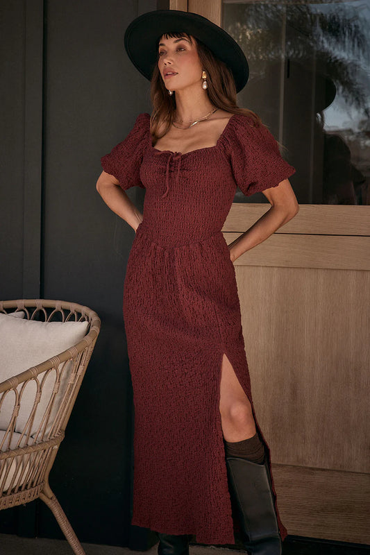 Woman in a burgundy dress with puffed sleeves and a slit, standing indoors.
