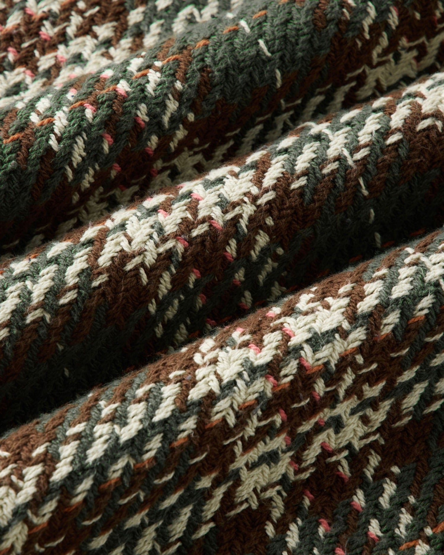 A close-up of a textured woven fabric with a houndstooth pattern in brown, dark green, cream, and small pink accents.
