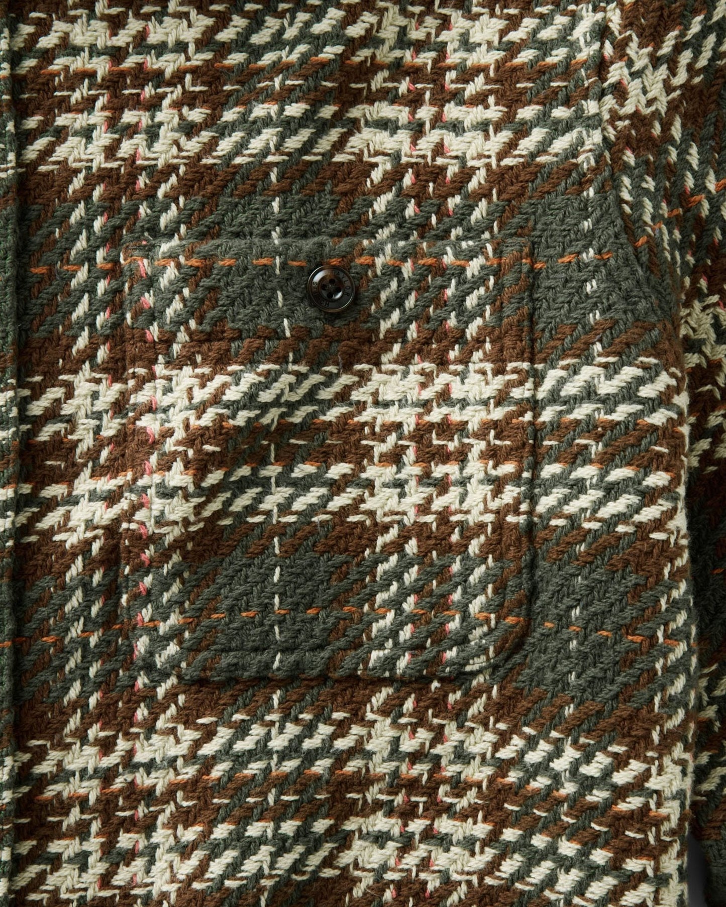 Close-up of a textured woven fabric with a brown, cream, and gray houndstooth pattern, featuring a buttoned pocket.