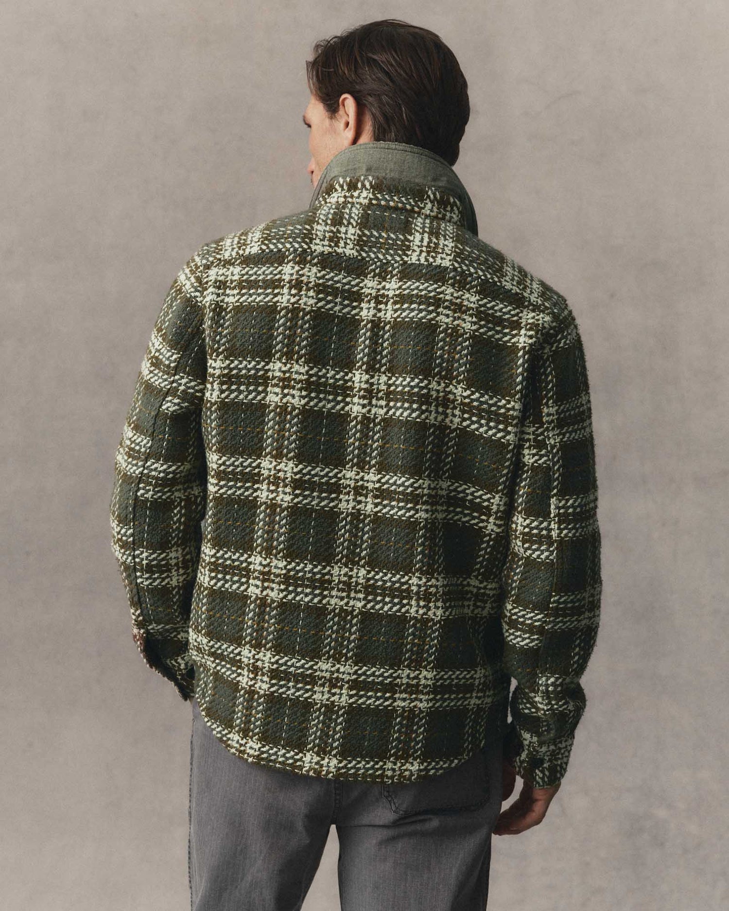 A man viewed from behind wearing a dark green and beige plaid overshirt with gray trousers, standing against a textured gray studio background.