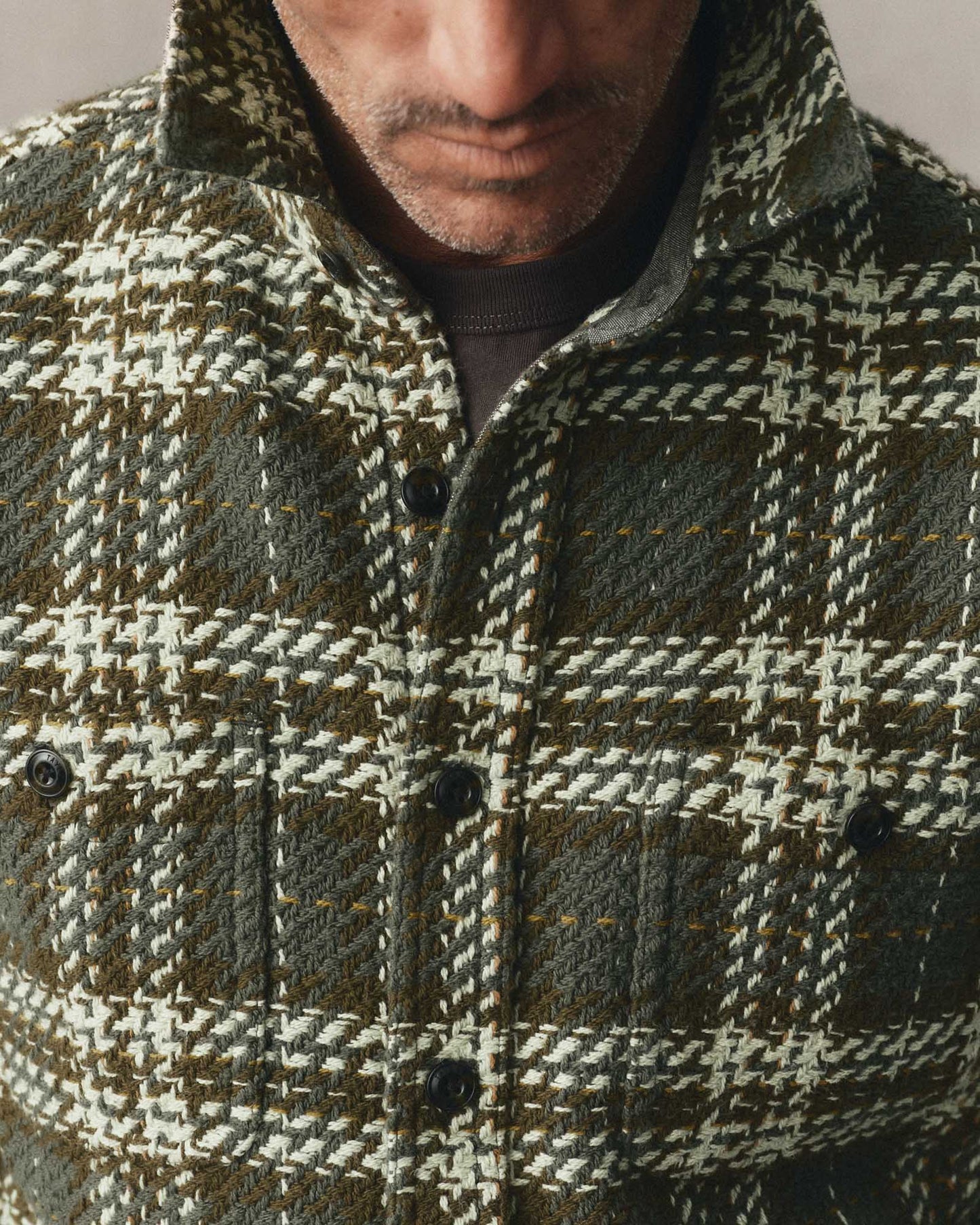 Close-up of a man wearing a textured plaid overshirt in olive green, beige, and brown tones, with a dark crew-neck undershirt visible at the collar.