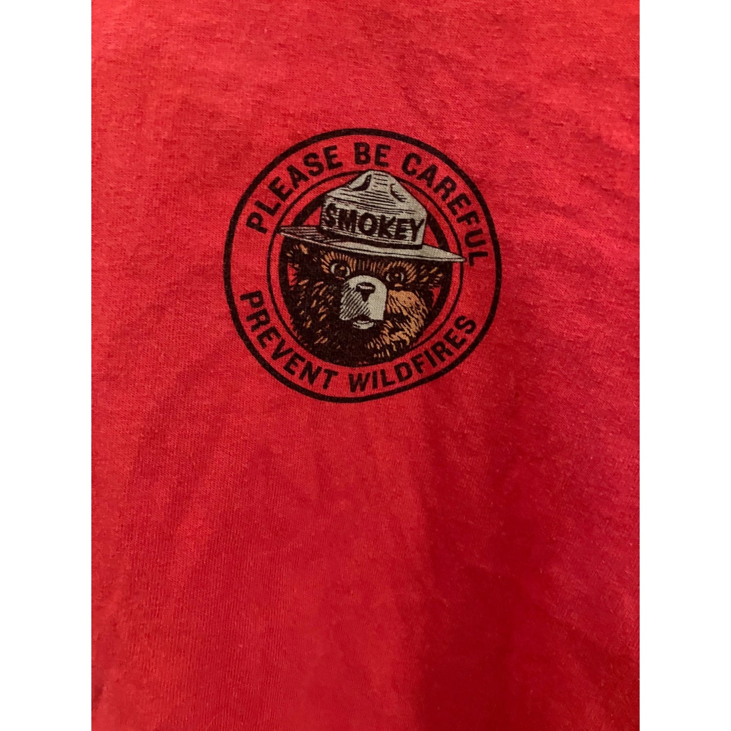 A close-up of a red t-shirt featuring a circular black graphic of the Smokey the Bear ranger hat and face, with the text 'PLEASE BE CAREFUL' and 'PREVENT WILDFIRES'.