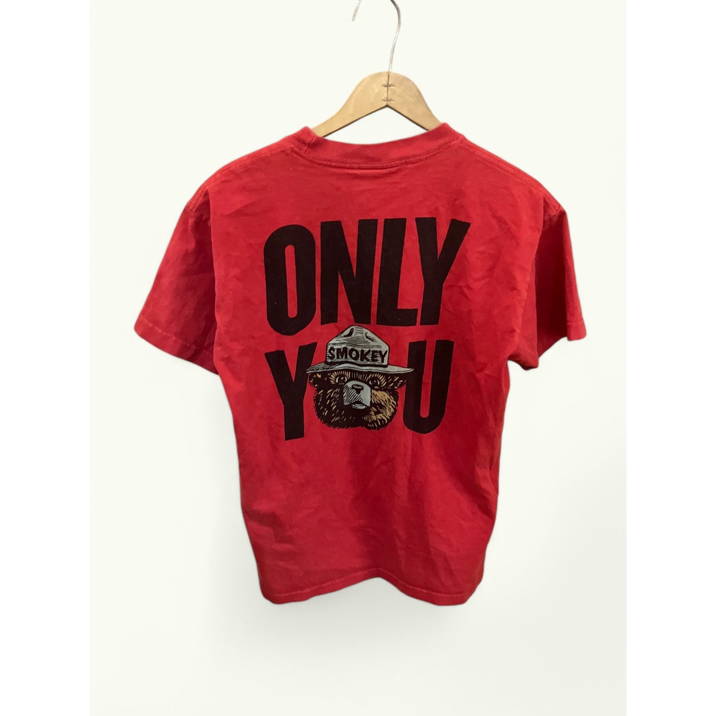 A red short-sleeve t-shirt hanging on a wooden hanger against a plain off-white wall. The back of the shirt features a large black graphic print reading 'ONLY YOU' with a circular 'Smokey' logo integrated into the lettering.