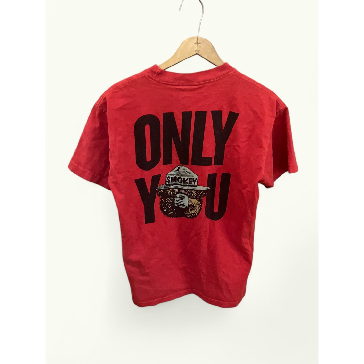 A red short-sleeve t-shirt hanging on a wooden hanger against a plain off-white wall. The back of the shirt features a large black graphic print reading 'ONLY YOU' with a circular 'Smokey' logo integrated into the lettering.