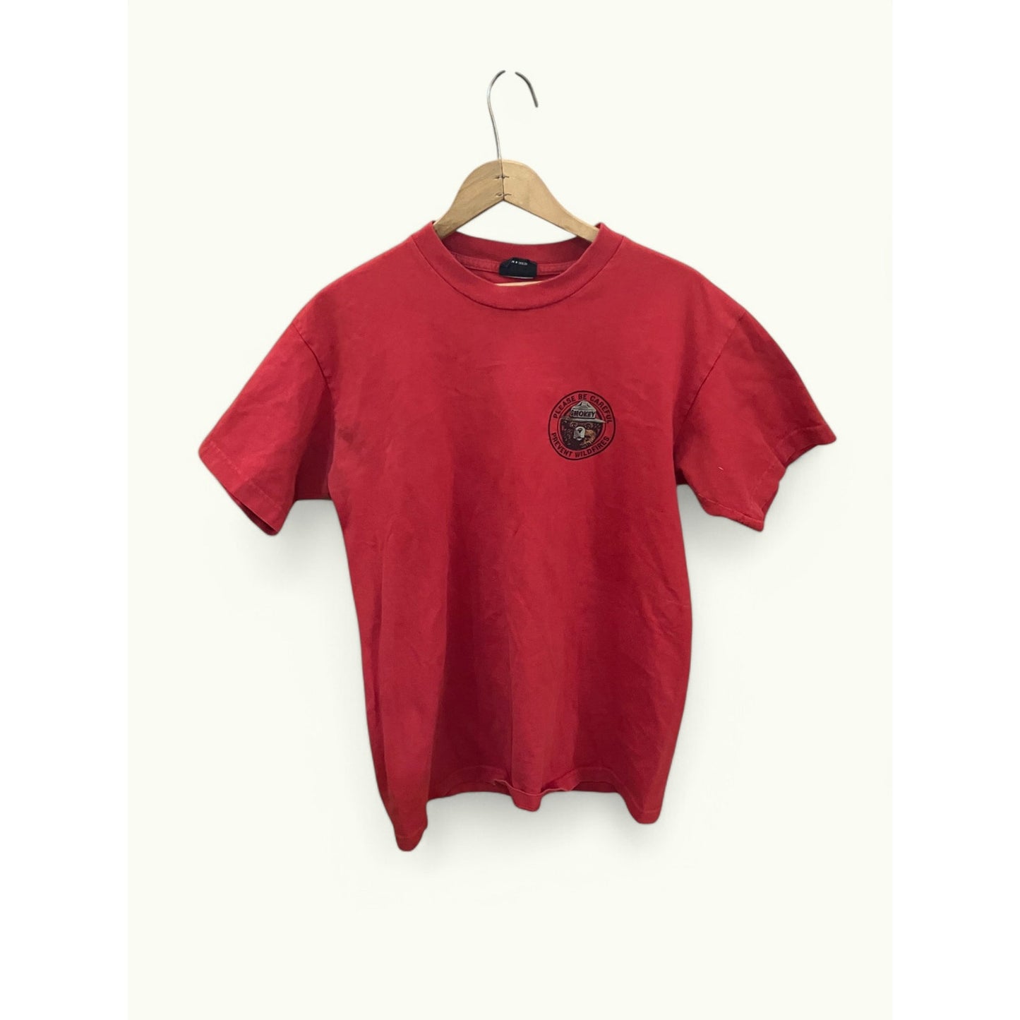 A red short-sleeve crewneck t-shirt hanging on a wooden hanger against a plain off-white background. The shirt features a circular black graphic printed on the left chest.