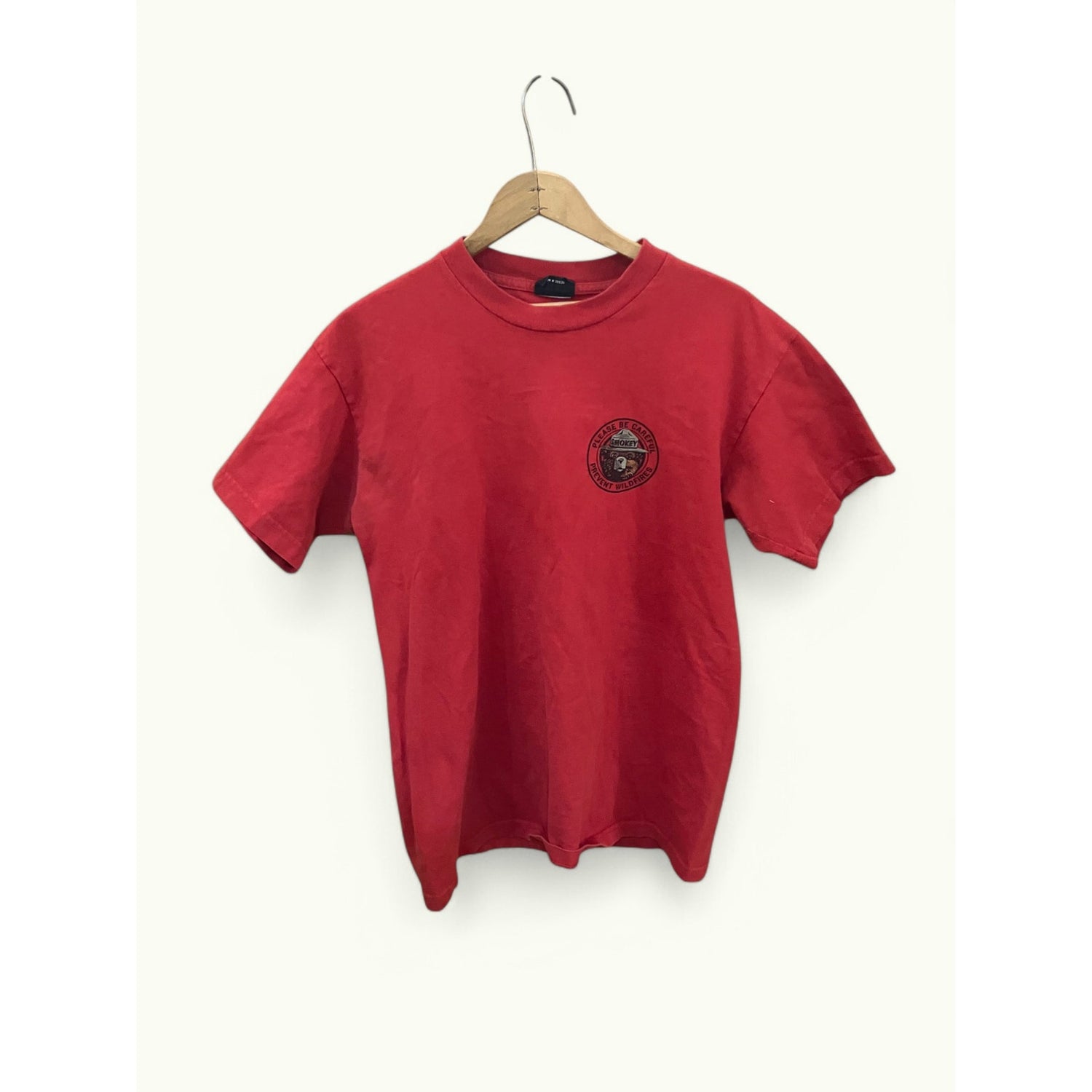 A red short-sleeve crewneck t-shirt hanging on a wooden hanger against a plain off-white background. The shirt features a circular black graphic printed on the left chest.