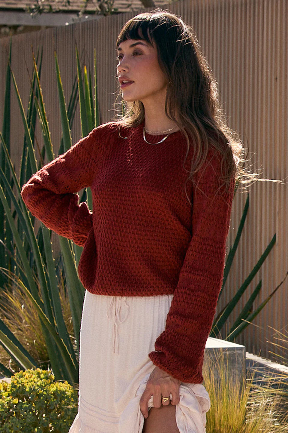Woman wearing a red sweater and white pants standing outdoors with plants in the background