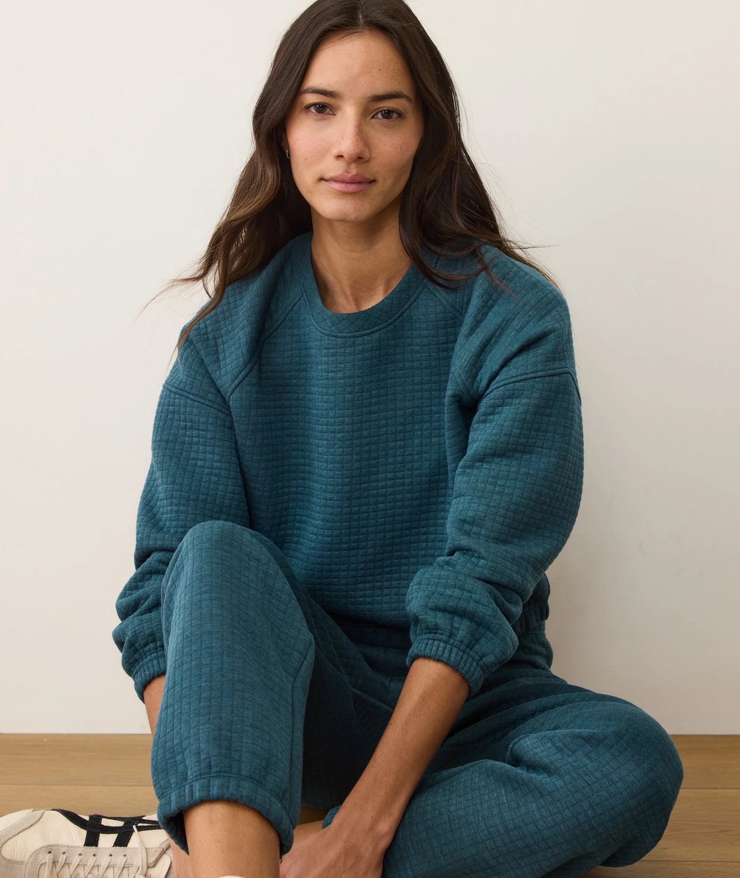 Woman wearing a teal sweater and pants sitting on the floor against a white wall.