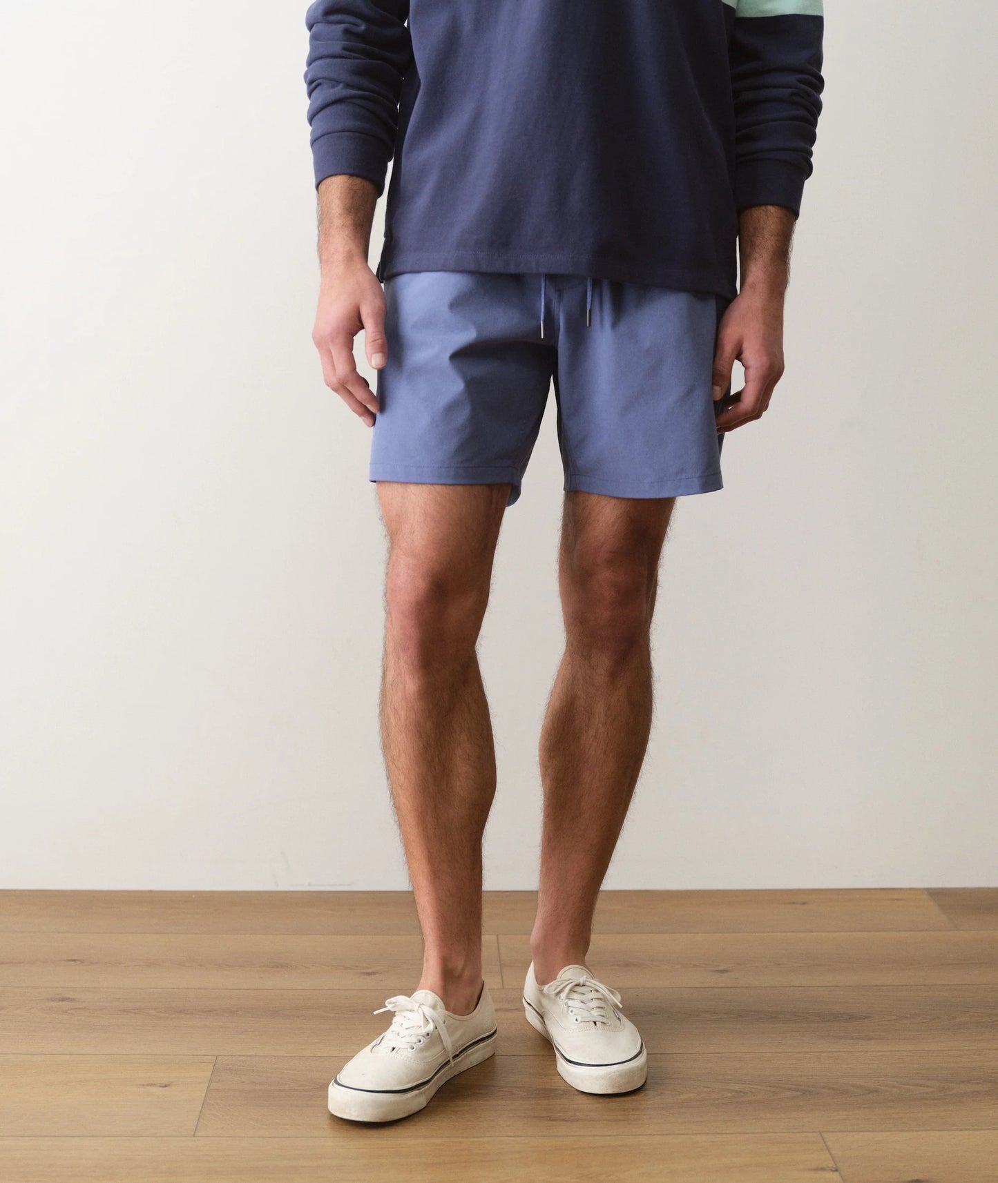 Person wearing blue shorts and a navy sweatshirt on a wooden floor with a white wall background
