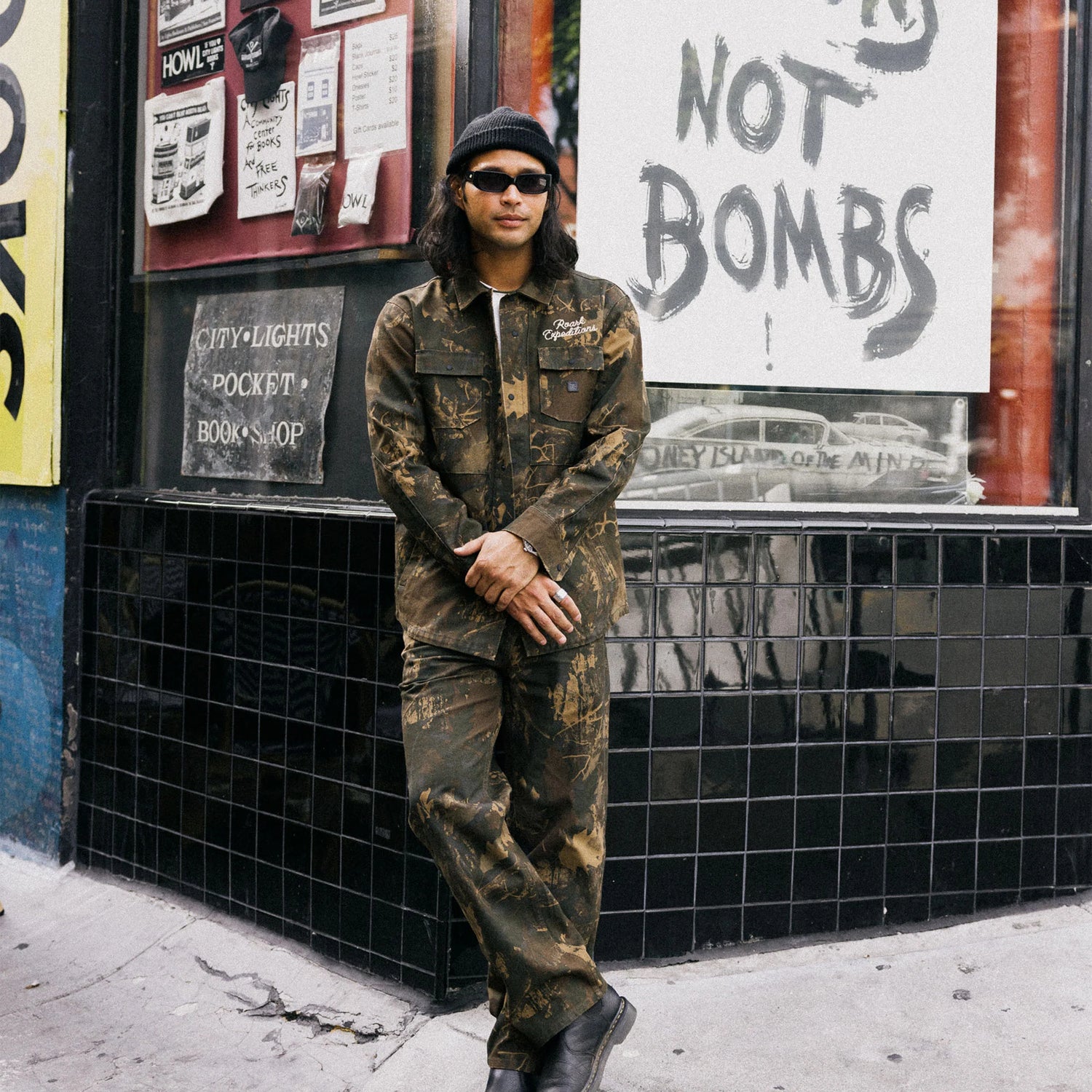 Person in camouflage outfit standing against a wall with posters and signs.