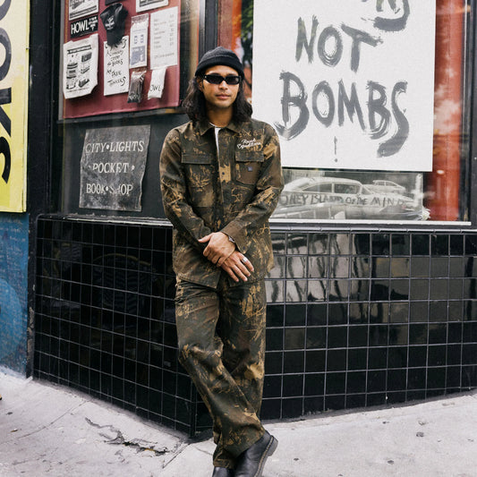 Person in camouflage outfit standing against a wall with posters and signs.