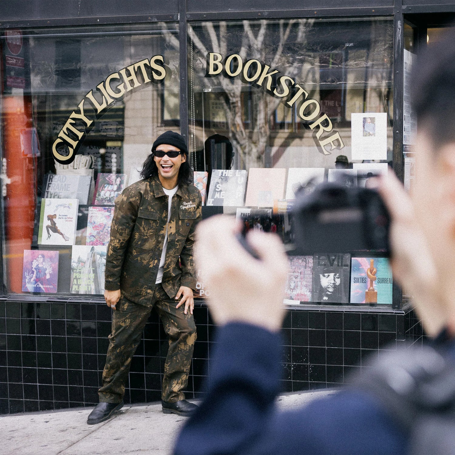 Person in camouflage outfit being photographed in front of 'City Lights Bookstore'.