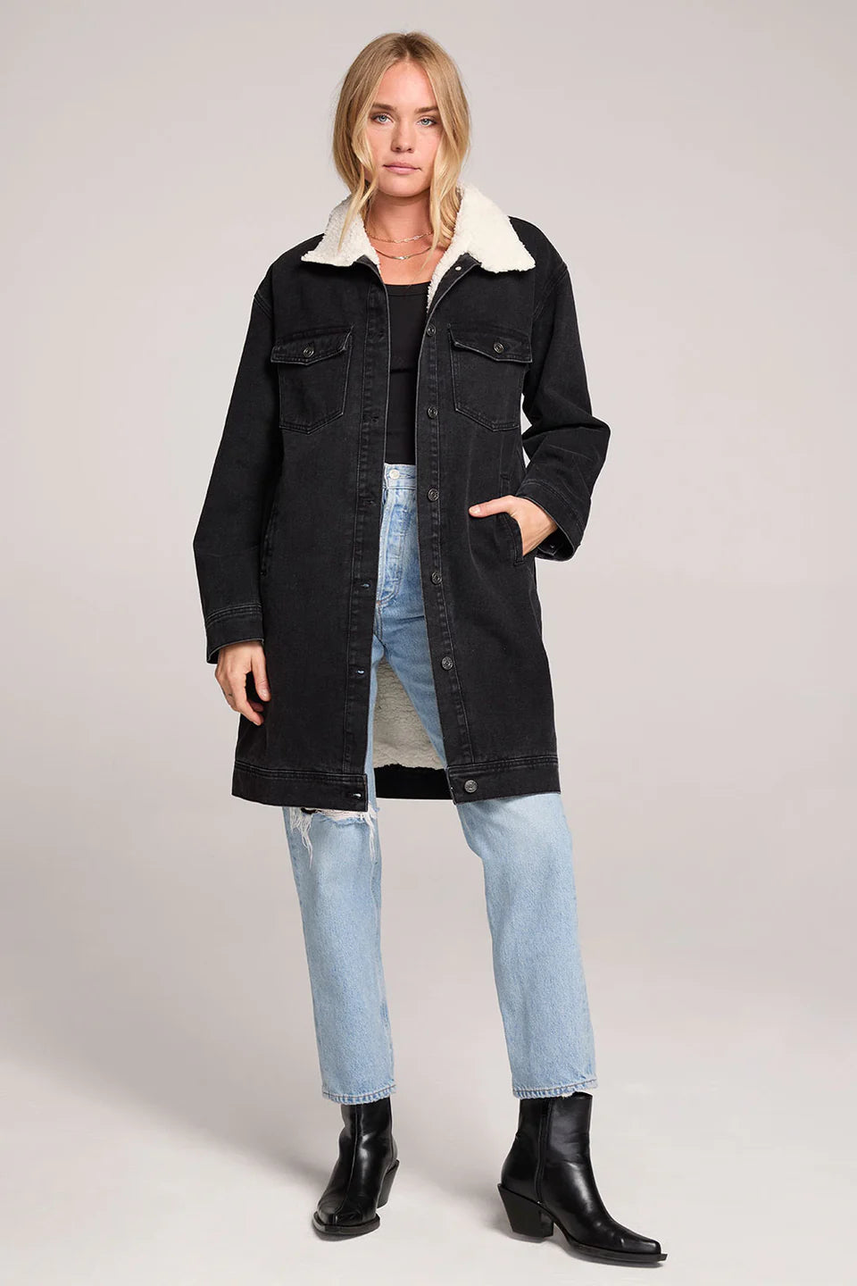 Woman wearing a black denim jacket with white fur collar, light blue jeans, and black boots on a plain background