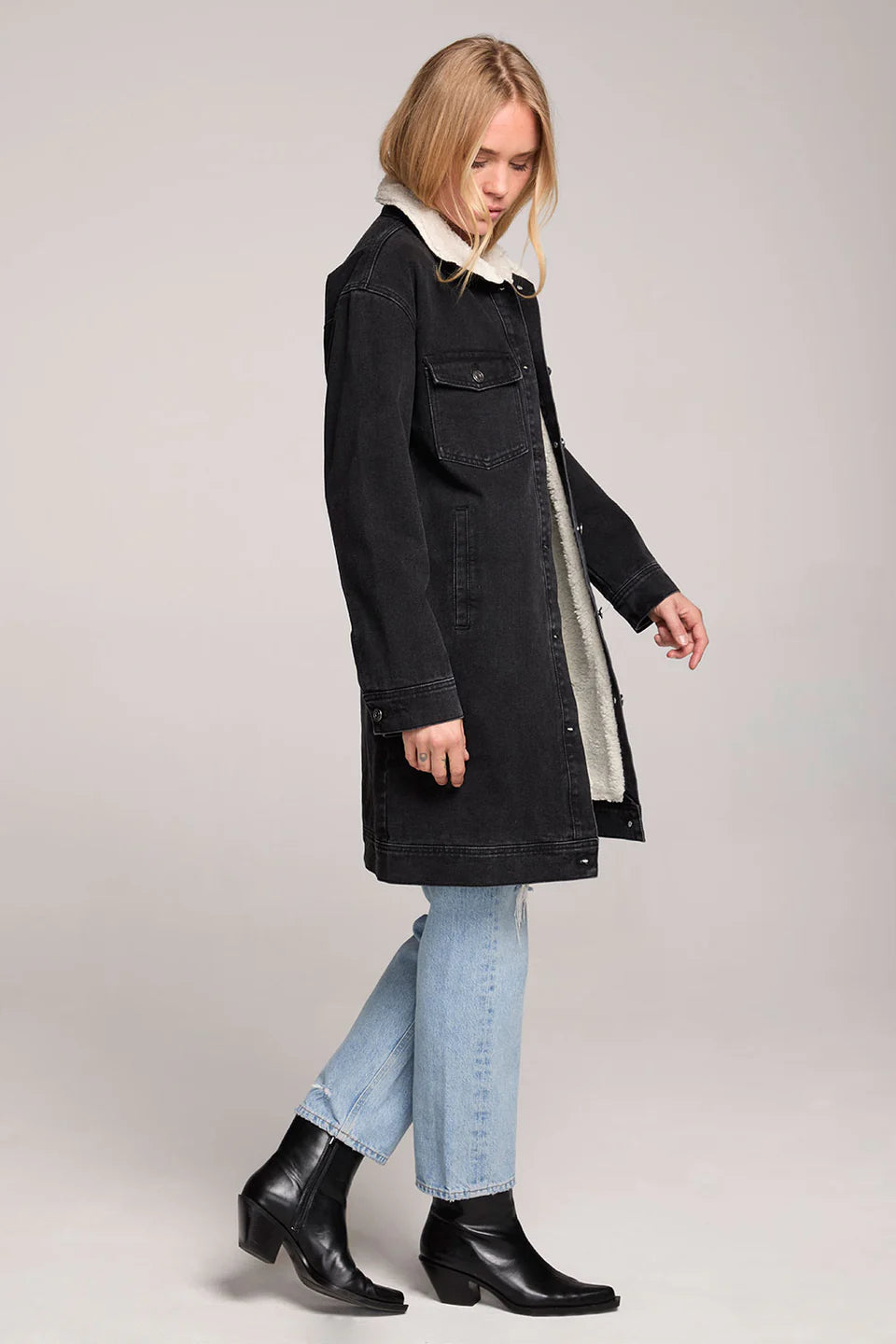 Woman wearing a black coat with white lining, light blue jeans, and black boots on a plain background