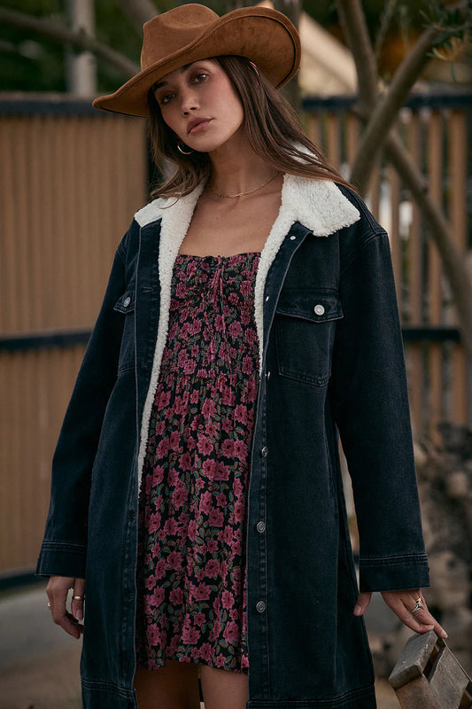 Woman wearing a floral dress under a dark denim coat with a fur-lined hood, standing outdoors.