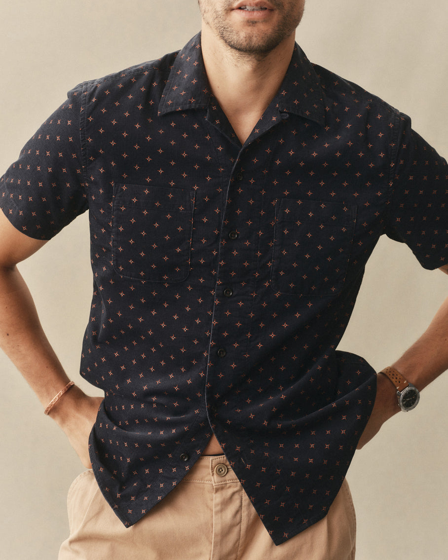 Man wearing a dark patterned shirt with beige pants against a neutral background
