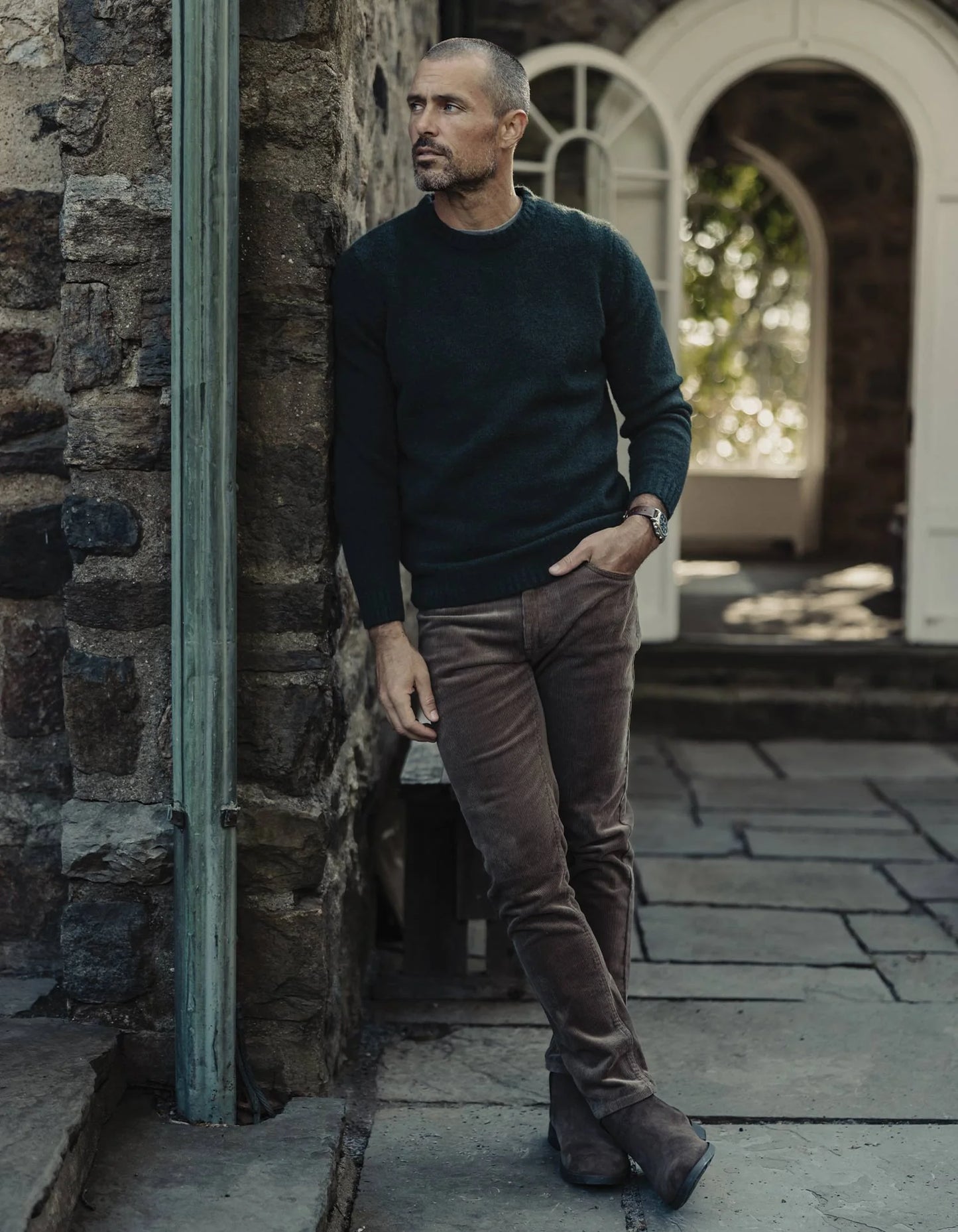 Man in a dark sweater and brown pants standing against a stone wall.
