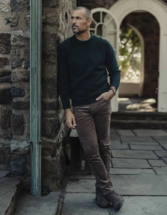 Man in a dark sweater and brown pants standing against a stone wall.