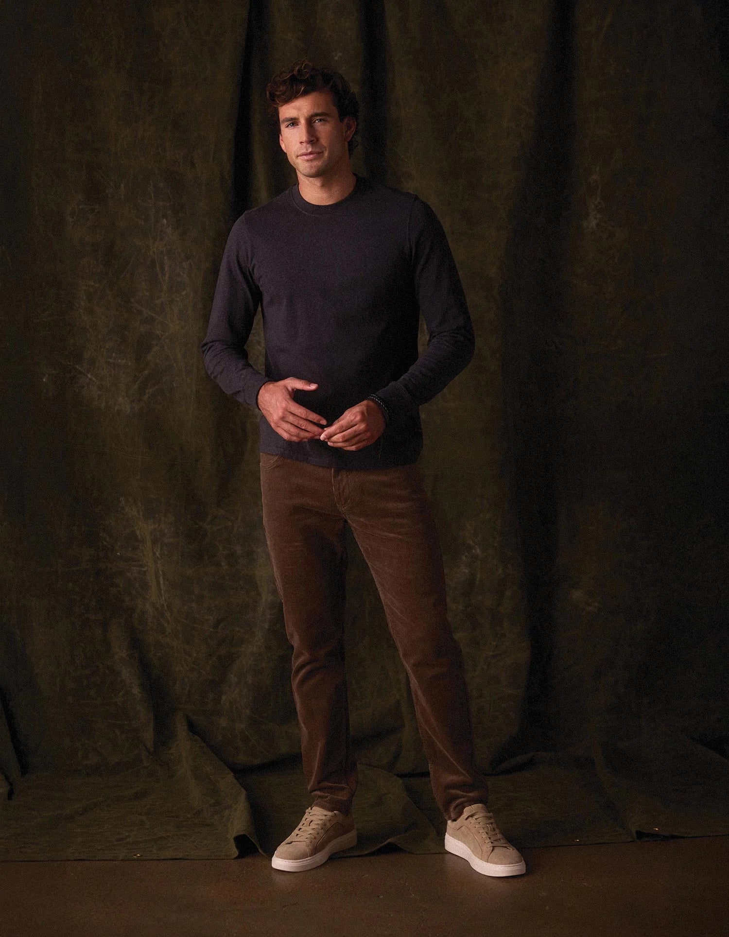 Man wearing a dark sweater and brown pants standing against a textured dark green curtain.