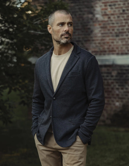 Man wearing a dark blazer over a beige shirt and pants, standing outdoors with a brick wall and tree in the background.