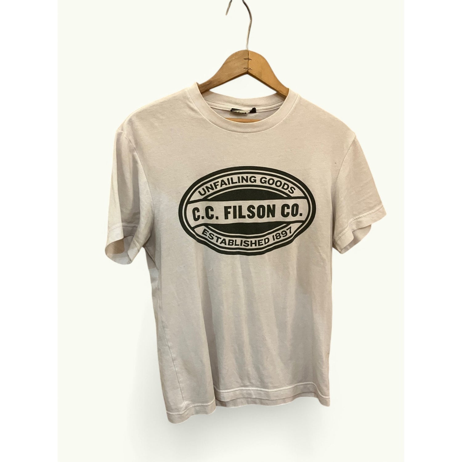 A light beige short-sleeve t-shirt hanging on a wooden hanger against a white wall. The shirt features a large black graphic print on the front with the text 'UNFAILING GOODS C.