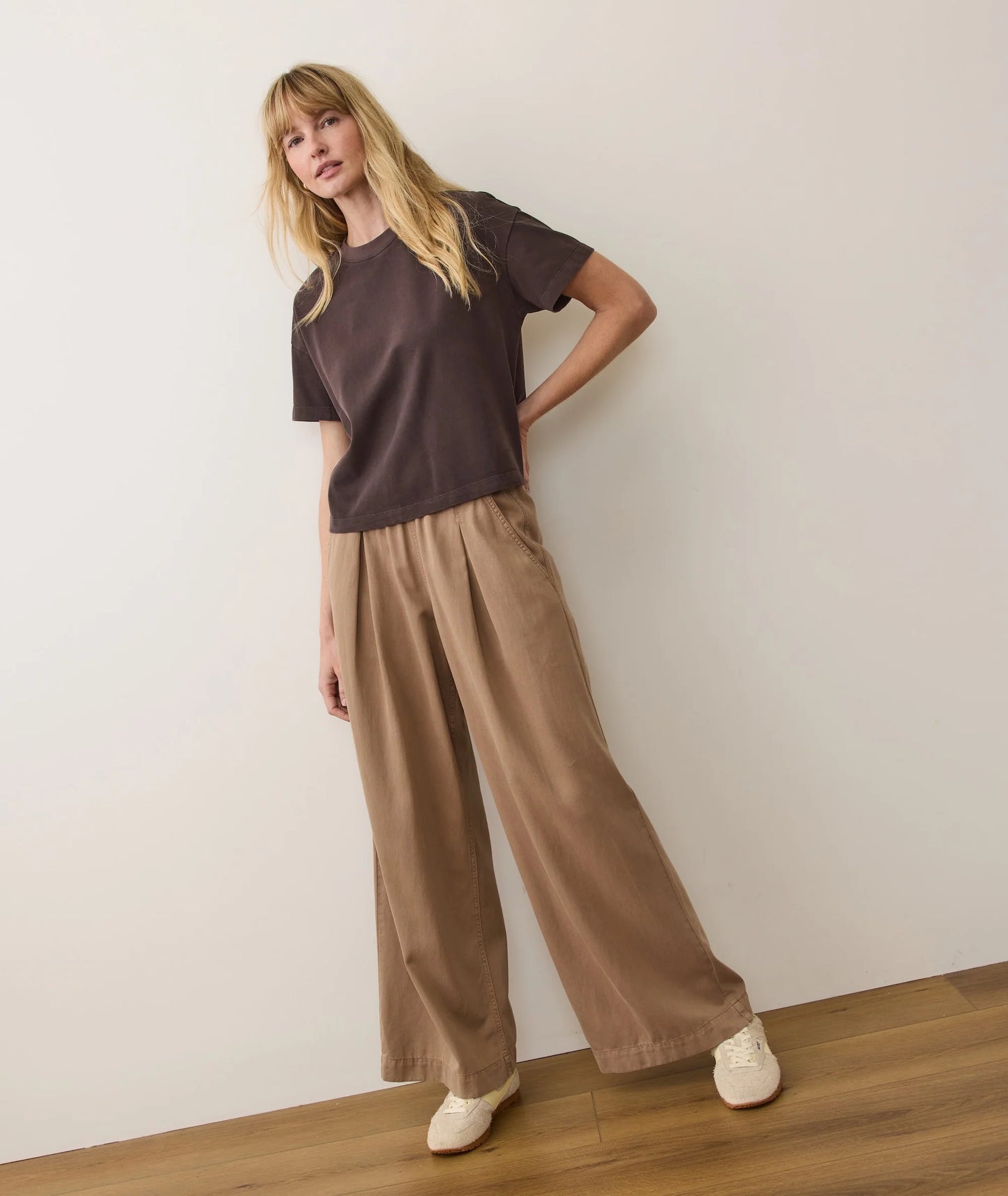Woman wearing a brown t-shirt and beige wide-leg pants against a white wall.