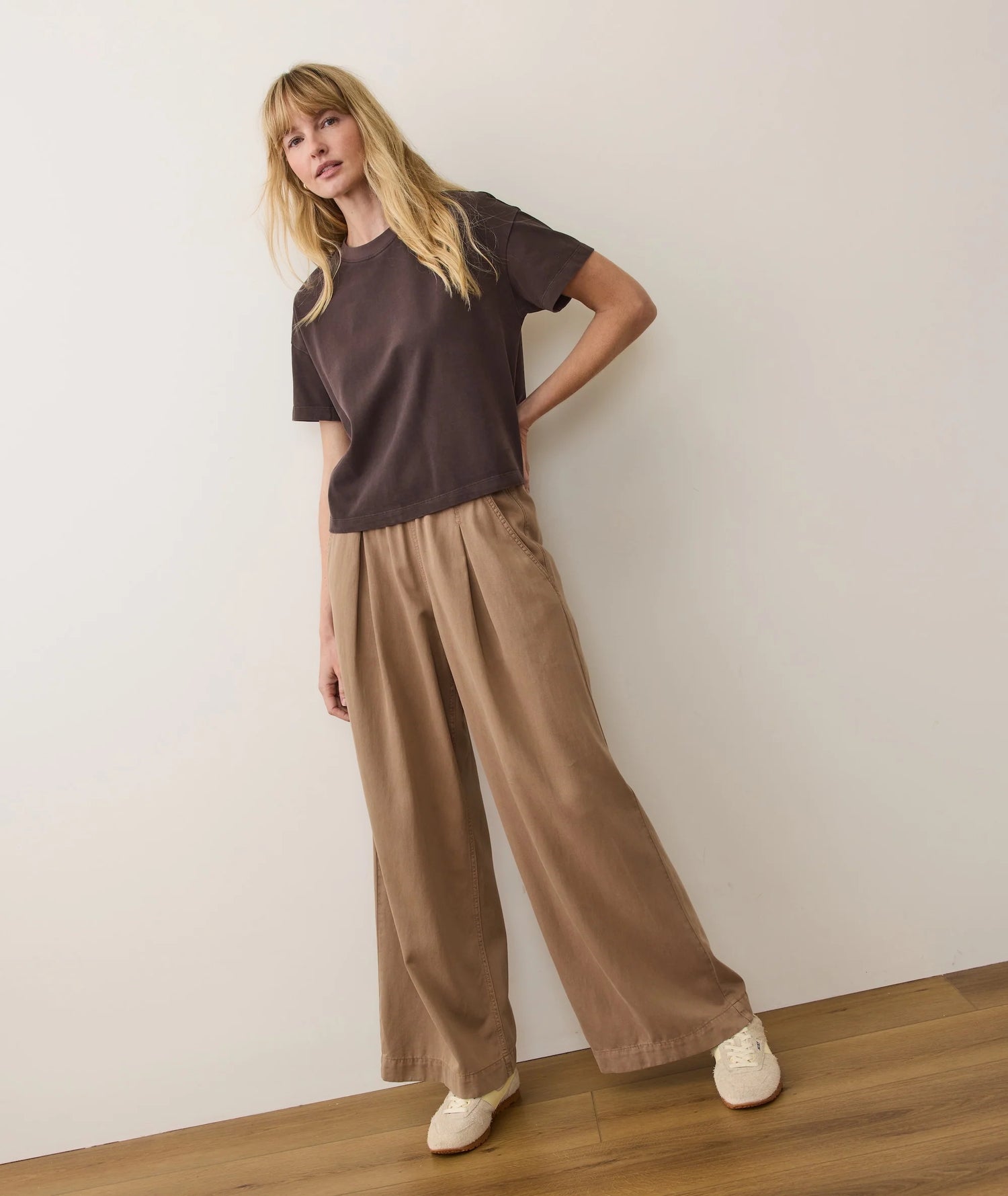 Woman wearing a brown t-shirt and beige wide-leg pants against a white wall.